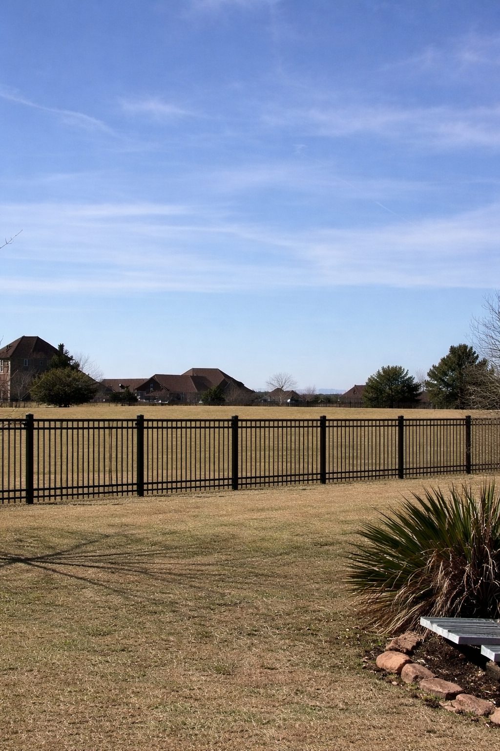 Black ornamental iron fence bordering an open field with homes in the distance